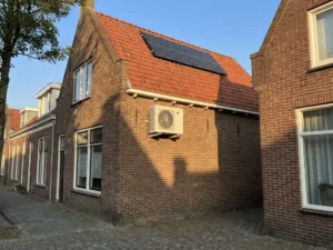 Witte airco-unit gemonteerd op bakstenen muur met zonnepanelen op rode dakpannen in Nederlandse woonwijk