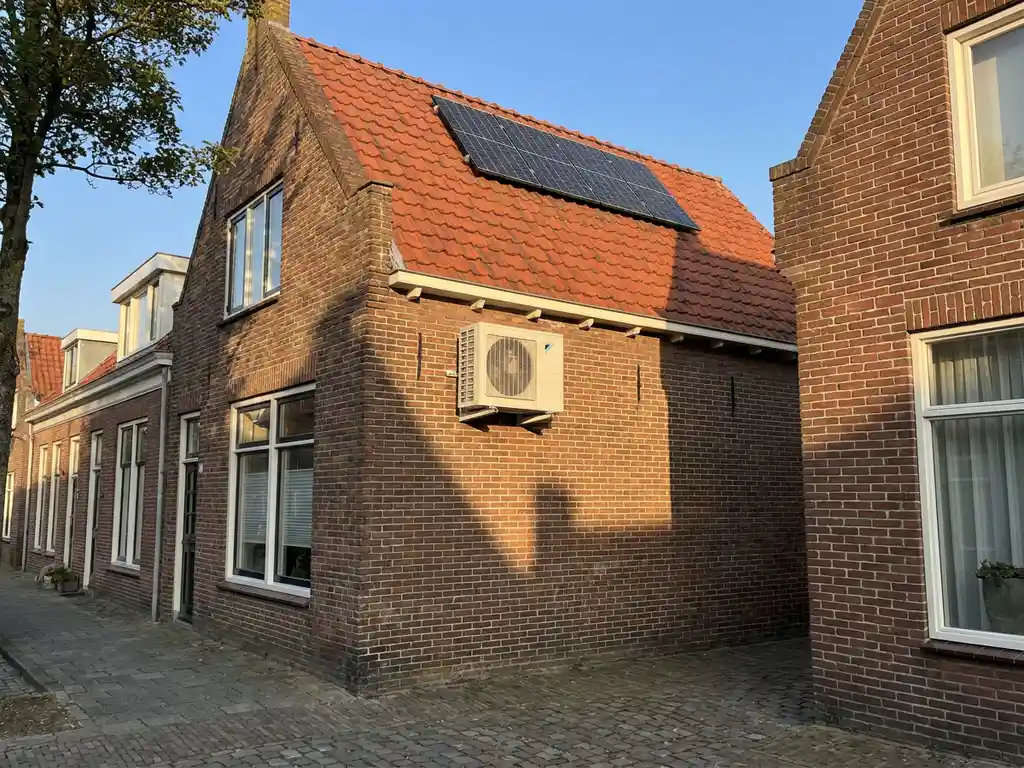 Witte airco-unit gemonteerd op bakstenen muur met zonnepanelen op rode dakpannen in Nederlandse woonwijk