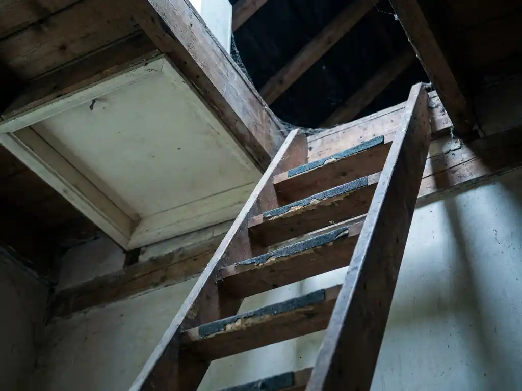 Steile houten vlizotrapladder tegen een wit dakluik, versleten antislipstrips, met zicht omhoog naar een stoffige zolderopening.