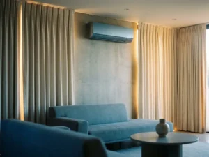 Moderne woonkamer met gesloten gordijnen en wandmonteerde airconditioning die zomerhitte weert, koele blauwe schaduwen met warme amberaccenten.